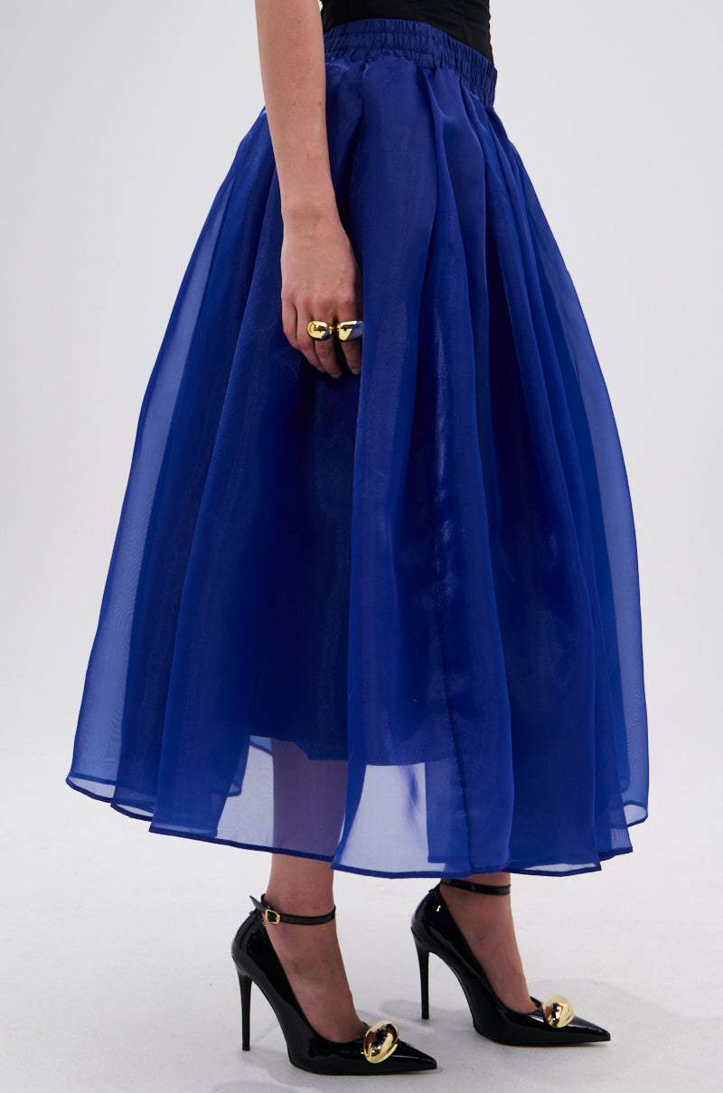 A person wears the STAY CLOSE TO ME MIDI SKIRT IN BLUE, a sheer royal blue A-line tulle skirt, paired with black high heels with gold accents and gold rings. The background is plain and light-colored. 2