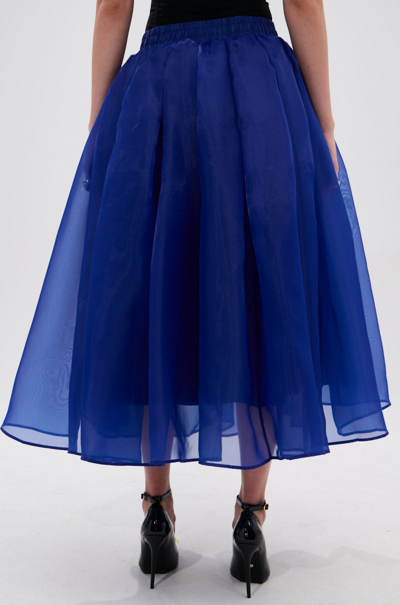 A person wears the STAY CLOSE TO ME MIDI SKIRT IN BLUE, an A-line, pleated royal blue midi skirt with a sheer chiffon overlay, paired with black ankle-strap heels. The back view highlights the skirt's flowy fabric and hemline. 3