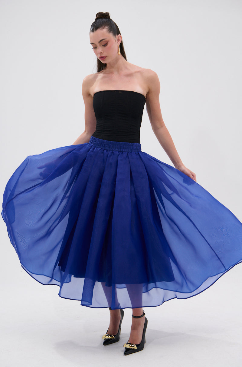 A woman stands with her skirt spread out, looking down. She wears the STAY CLOSE TO ME MIDI SKIRT IN BLUE with a black strapless top, black heels, and has long dark hair tied back. 4