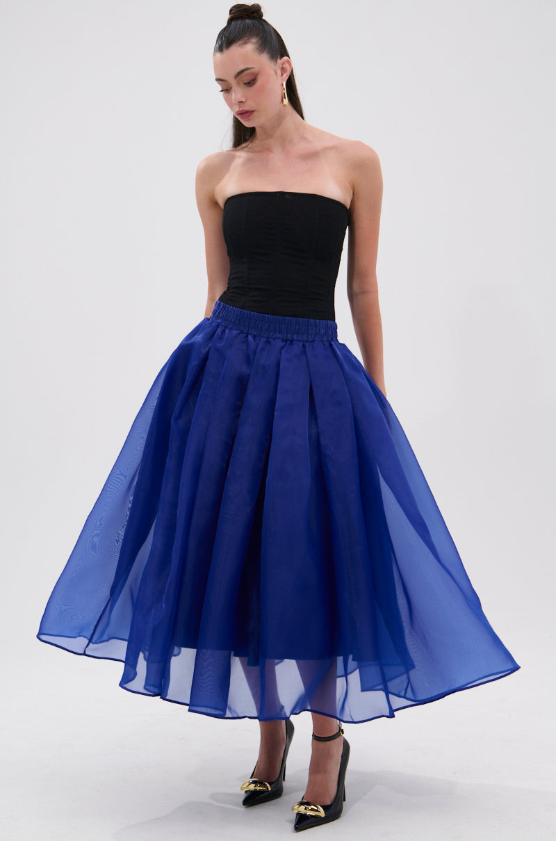 A woman wears the STAY CLOSE TO ME MIDI SKIRT IN BLUE with a black strapless top and black heels with gold accents, standing against a plain white background, looking down with her hands by her sides. 5
