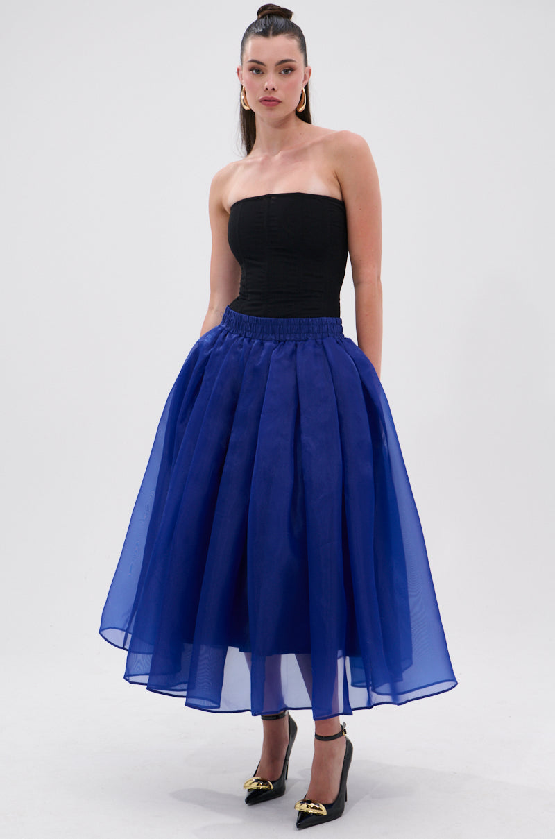 A woman poses against a plain background wearing the STAY CLOSE TO ME MIDI SKIRT IN BLUE, featuring a high-waisted, sheer chiffon A-line silhouette. She completes her look with a black strapless top, gold earrings, and black heels. 6