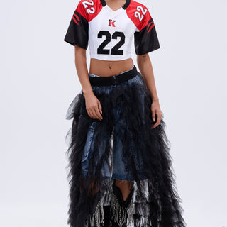 A woman wears the DRESSED TO IMPRESS MAXI SKIRT, a layered black tulle mesh piece over denim, paired with a cropped sports jersey (#22), black pointed fringe boots, hoop earrings, and tied-back hair. The background is plain white.