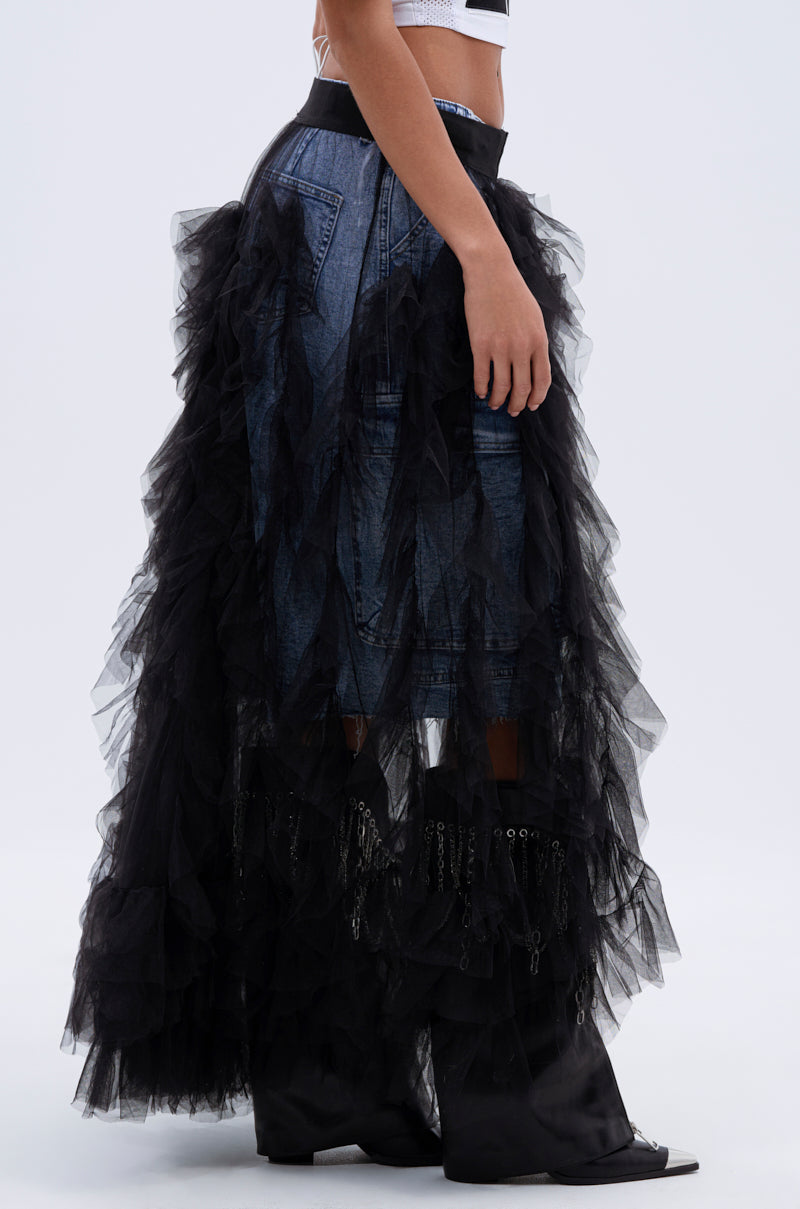 Someone wears the DRESSED TO IMPRESS MAXI SKIRT—high-rise denim with black ruffled tulle—paired with matching black boots, shown in profile against a plain light background.