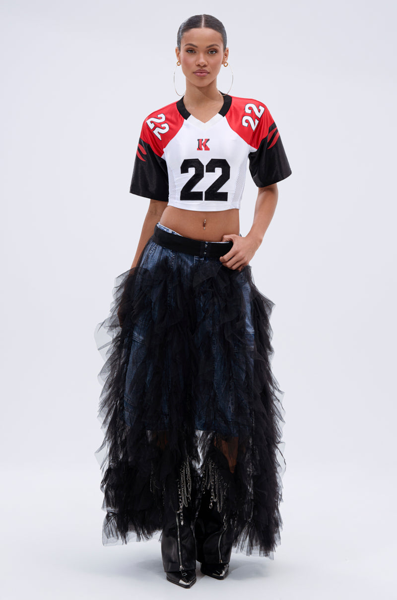 A woman poses against a plain backdrop in the DRESSED TO IMPRESS MAXI SKIRT, styled with a cropped football jersey featuring the number 22, shiny black boots, hoop earrings, and her hair pulled back.