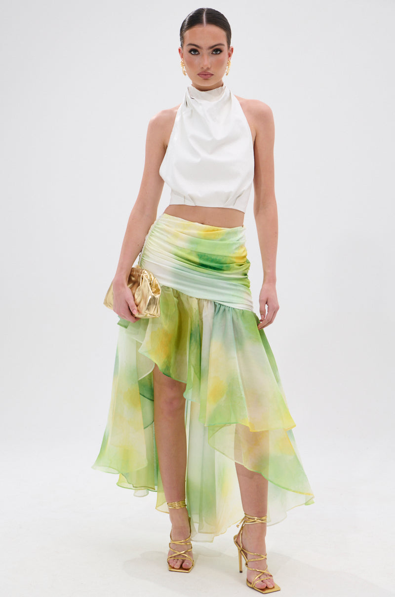 A woman poses in a sleeveless white halter top and the LIMONE CHIFFON MIDI SKIRT, featuring a green-yellow watercolor print and asymmetrical silhouette. She completes the look with gold strappy heels, a gold clutch, bold makeup, and pulled-back hair. 0