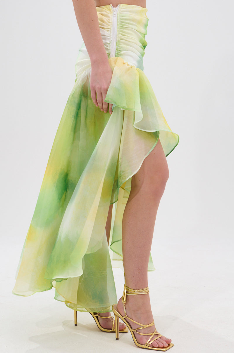 A person wearing the LIMONE CHIFFON MIDI SKIRT with green and yellow watercolor patterns, an asymmetrical silhouette, and a ruched waistband, paired with gold strappy high-heeled sandals, stands against a plain white background. 3