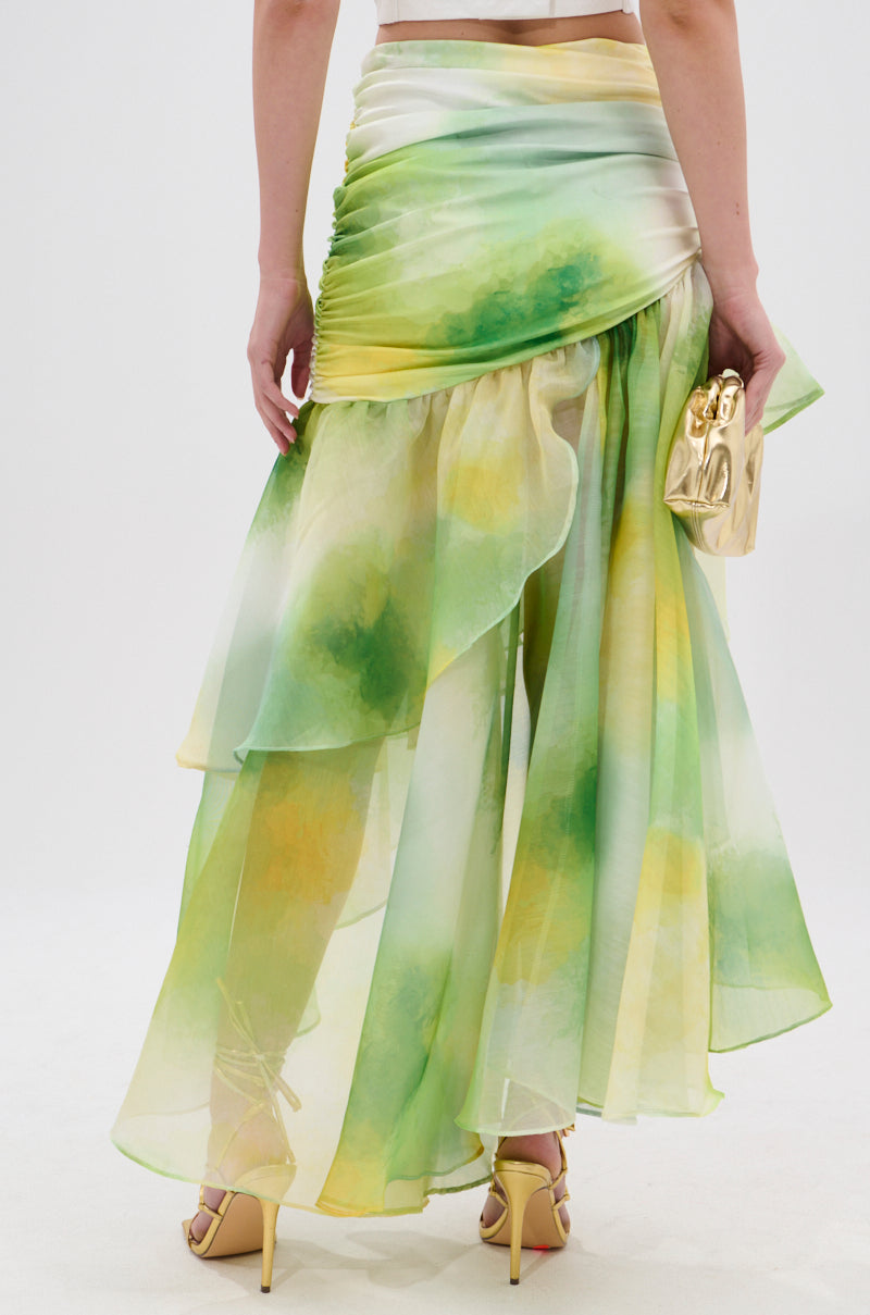 A person is shown from the waist down, wearing the LIMONE CHIFFON MIDI SKIRT in green and yellow with an asymmetrical silhouette, yellow high-heeled sandals, holding a shiny gold clutch. The background is plain white. 4