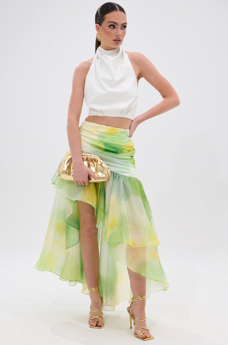 A woman poses against a white background in the LIMONE CHIFFON MIDI SKIRT, an asymmetrical green and yellow chiffon skirt with a ruched waistband. She pairs it with a white halter crop top, gold clutch, strappy heels, and slicked-back hair. 6