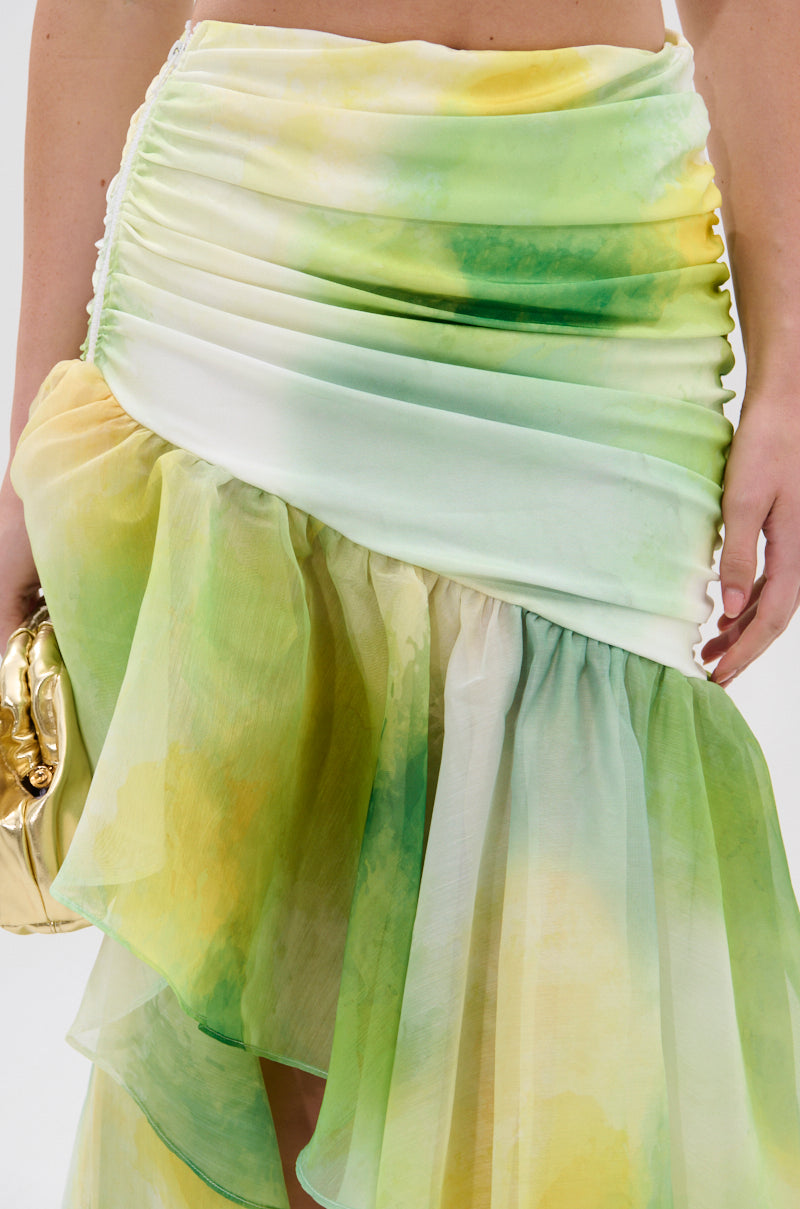 A close-up of a woman wearing the LIMONE CHIFFON MIDI SKIRT, featuring a ruched waistband, asymmetrical silhouette, and green, yellow, and white watercolor-style patterns, paired with a gold clutch. 7
