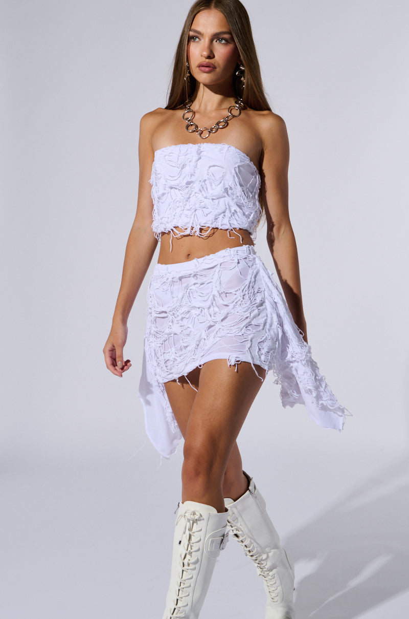 A woman with long hair walks confidently wearing a strapless white crop top and an asymmetrical skirt similar to the QUEEN OF LOVE DISTRESSED MINI SKIRT IN WHITE, which features a textured, floral design. She is accessorized with a chunky necklace and white lace-up knee-high boots. The background is plain and well-lit.