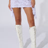 A model stands against a white background wearing the QUEEN OF LOVE DISTRESSED MINI SKIRT IN WHITE, featuring an asymmetrical silhouette and high-waisted design. Her legs are adorned with knee-high white lace-up heeled boots, creating a delicate yet edgy style.