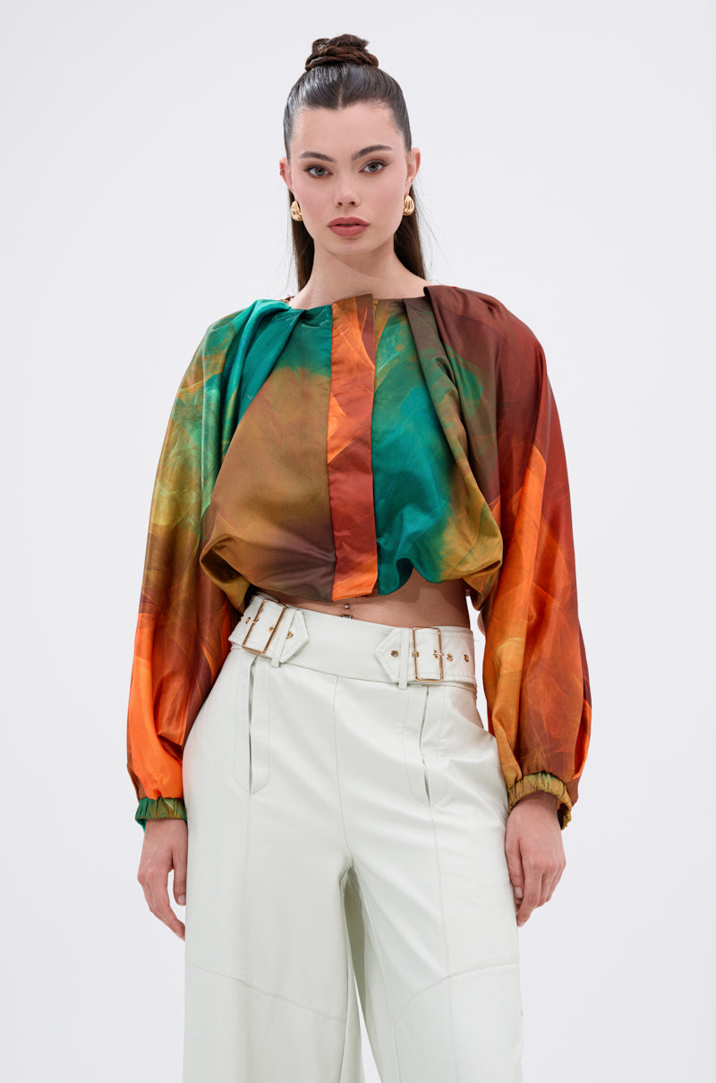 A woman with a high bun models the TIFFANY OMBRE BLOUSE, showcasing its green, orange, and brown ombré print with balloon sleeves. She pairs it with high-waisted white pants and a matching belt against a plain background.