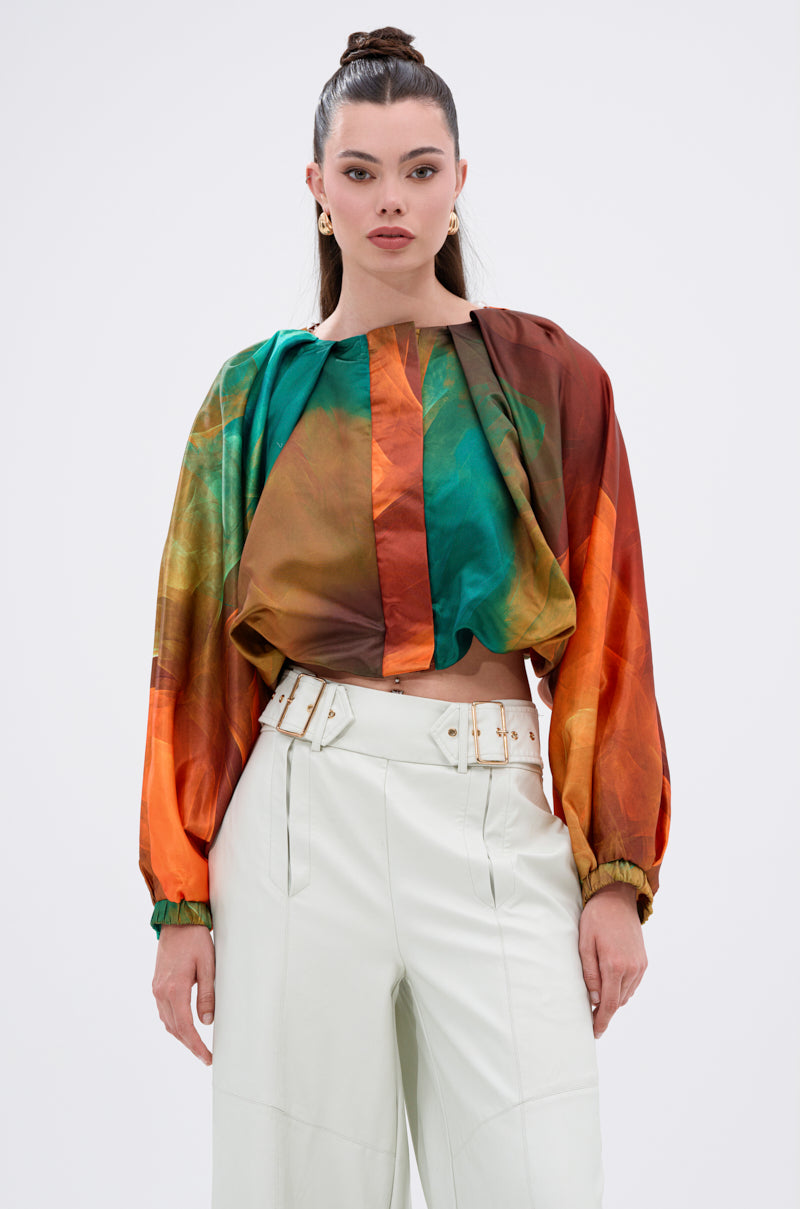 A woman with long dark hair in a high ponytail wears the TIFFANY OMBRE BLOUSE, featuring abstract orange, green, and brown patterns with balloon sleeves, paired with high-waisted white pants and a wide belt against a light background.