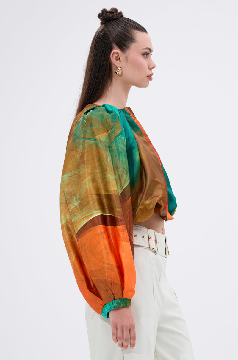 A woman with long brown hair in a half-up topknot models the TIFFANY OMBRE BLOUSE with balloon sleeves, white high-waisted pants, and gold earrings, standing in profile against a plain background.