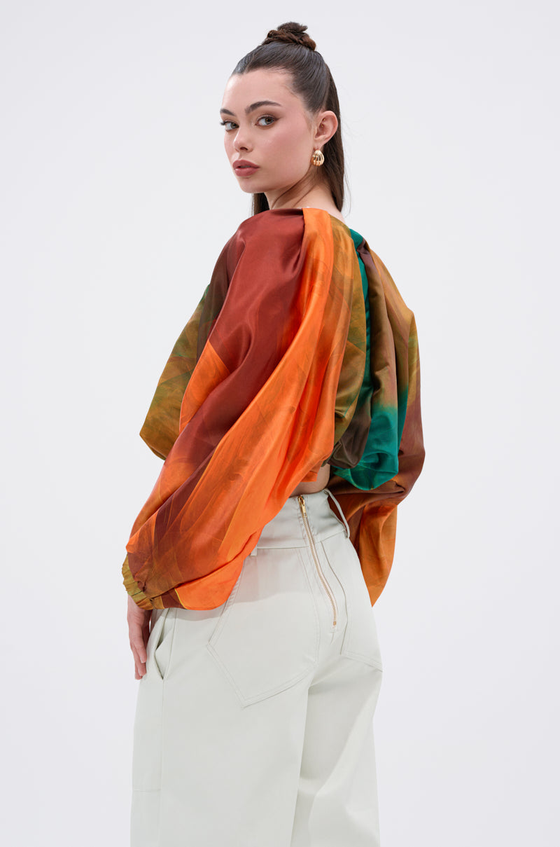 A woman with dark hair in a topknot wears the TIFFANY OMBRE BLOUSE, featuring billowy balloon sleeves, paired with high-waisted white pants and poses over her shoulder against a plain background.