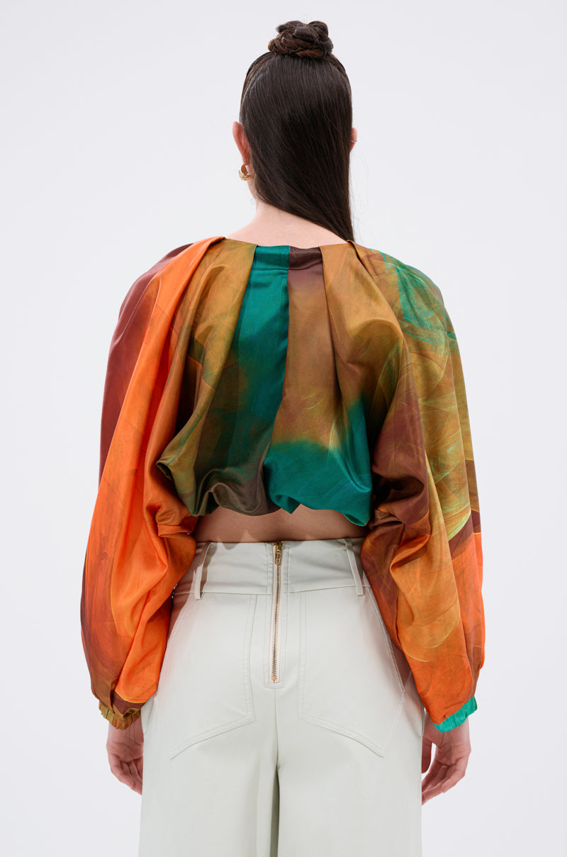 A person stands facing away, wearing the TIFFANY OMBRE BLOUSE with balloon sleeves in orange, green, and brown ombré. It's paired with high-waisted white pants with visible back seams and a center zipper; their hair is sleekly tied back.