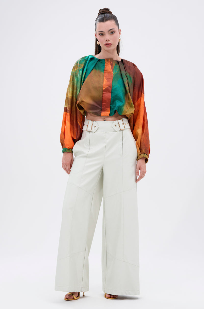 A woman stands confidently in the TIFFANY OMBRE BLOUSE with balloon sleeves, tucked into high-waisted, wide-leg white pants featuring a double-belted waistband. Her hair is tied back and she completes the look with heeled sandals.