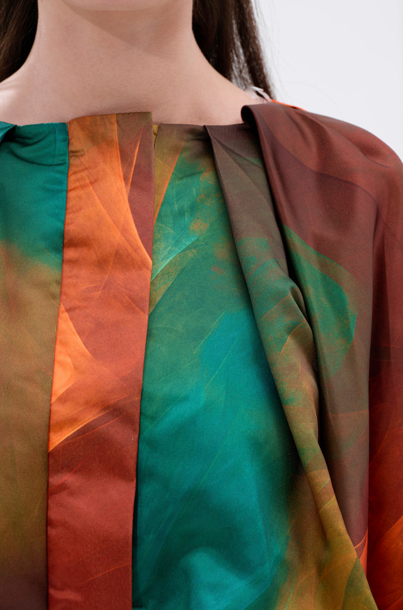 Close-up of a person wearing the TIFFANY OMBRE BLOUSE, featuring vibrant shades of orange, green, teal, and brown in an ombré print with pleats and folds. Only the lower half of the face and neck are visible.