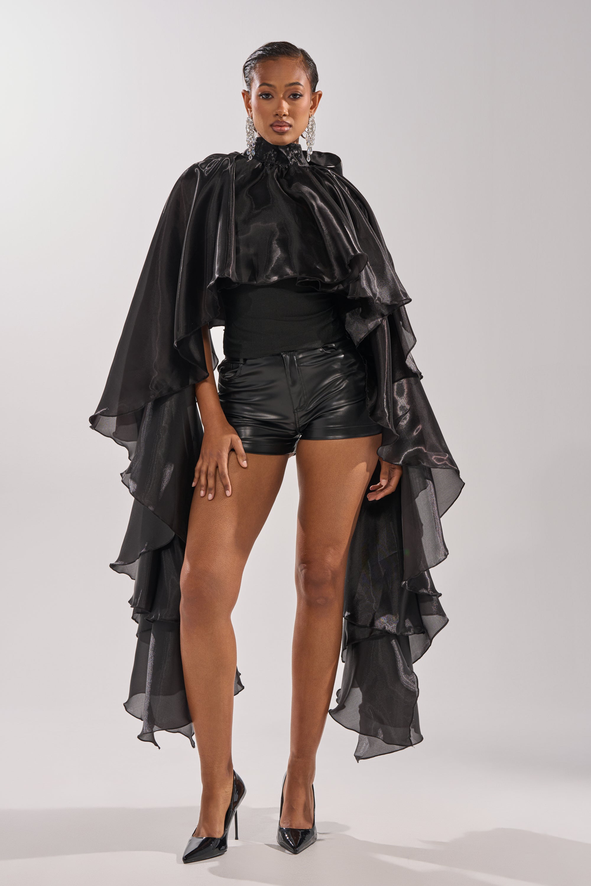 A woman stands confidently against a gray background wearing the COOLEST ON THE BLOCK BLOUSE, paired with black high-waisted shorts, a shiny black ruffle cape, pointed high heels, and statement earrings. 0