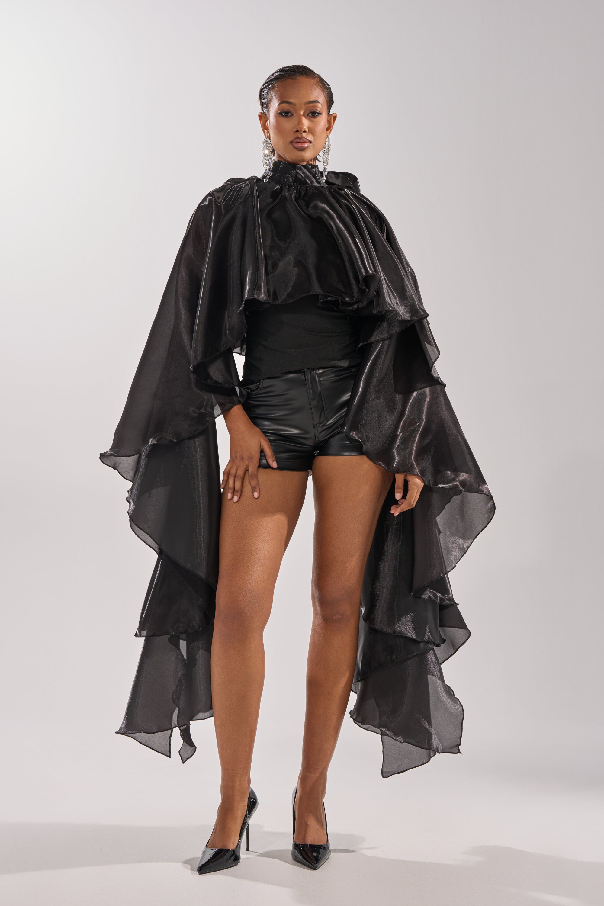 Wearing the COOLEST ON THE BLOCK BLOUSE, a woman exudes confidence in a dramatic ruffle top, black cape, high-neck shirt, and leather shorts. Black heels and slicked-back hair complete her bold, modern look. 1