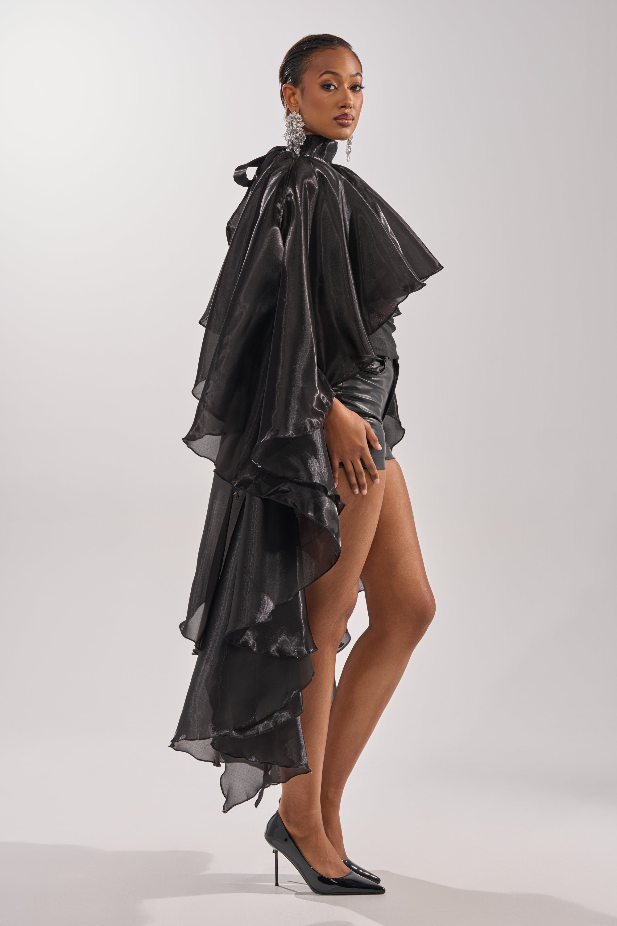 A woman models the COOLEST ON THE BLOCK BLOUSE, a dramatic black chiffon top with ruffles and a long train, styled with high-waisted shorts, statement earrings, and black heels against a plain light background. 2