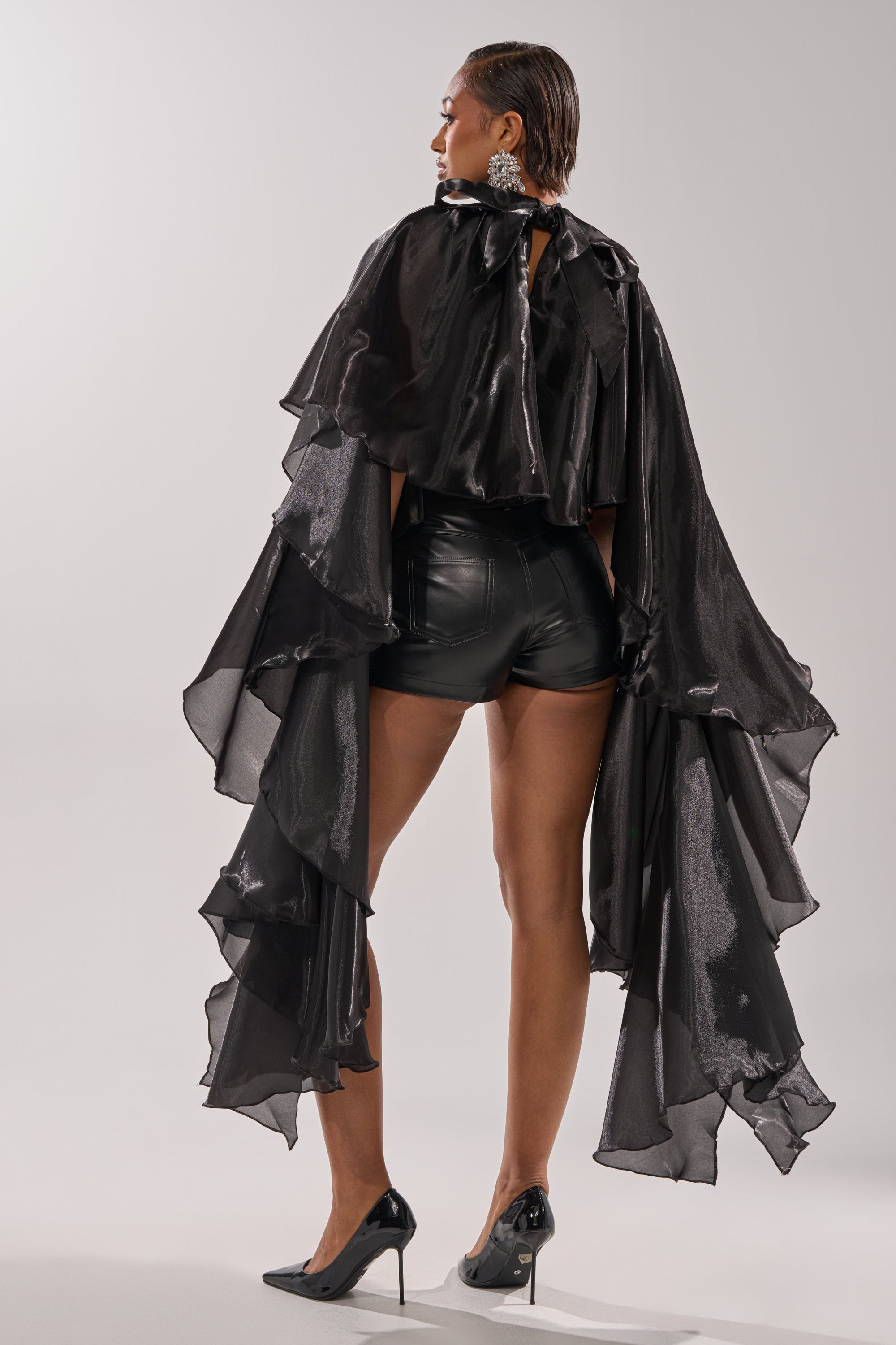 Dressed in the COOLEST ON THE BLOCK BLOUSE, a woman faces away in leather shorts and high heels, showcasing the dramatic ruffle design and sheer cape. Her slicked-back hair stands out against the plain, light background. 3