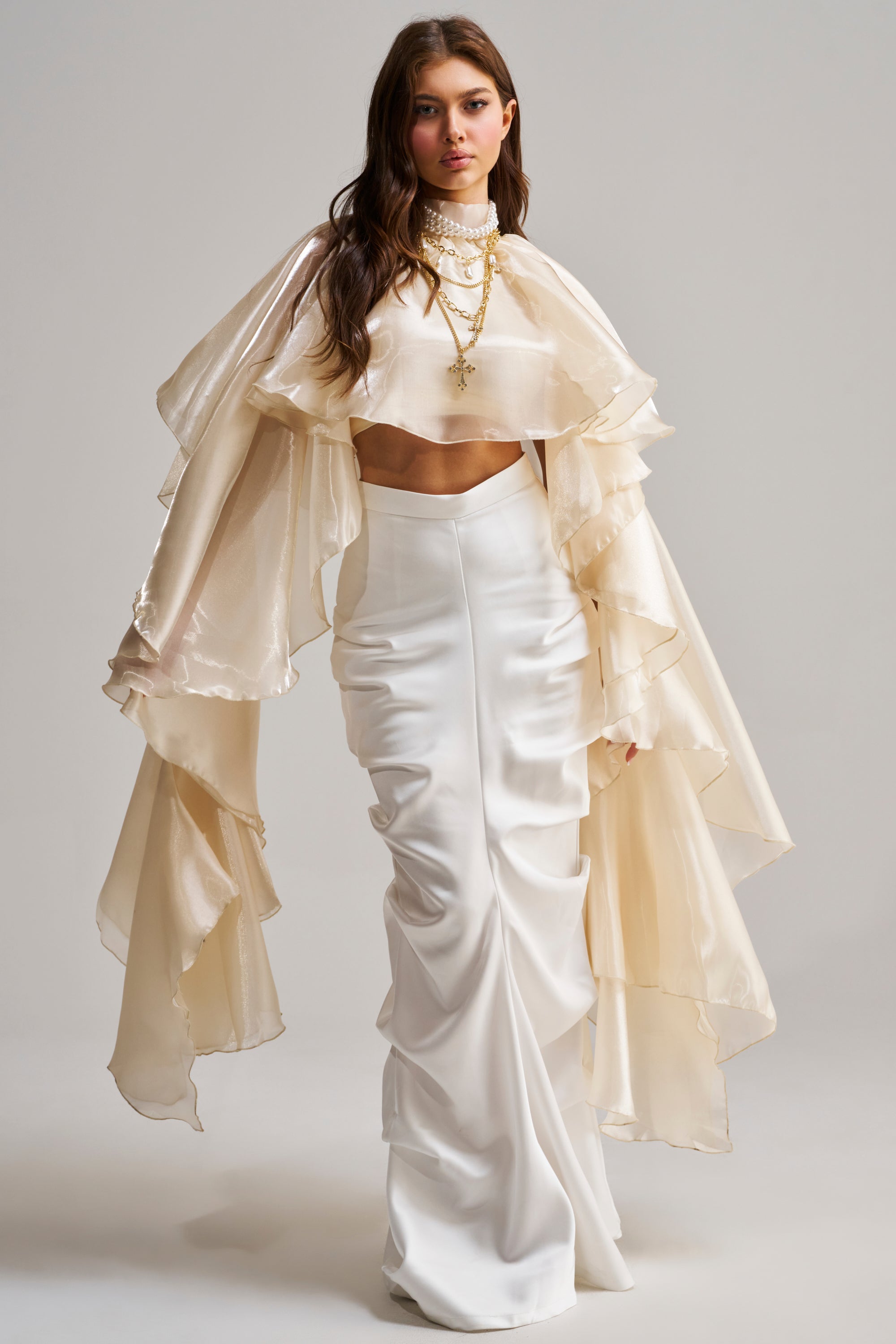 A woman wears the STAR OF THE NIGHT TOP, a couture-inspired cream crop top with dramatic layered sleeves, paired with a fitted white skirt and gold necklaces, standing against a plain light background.