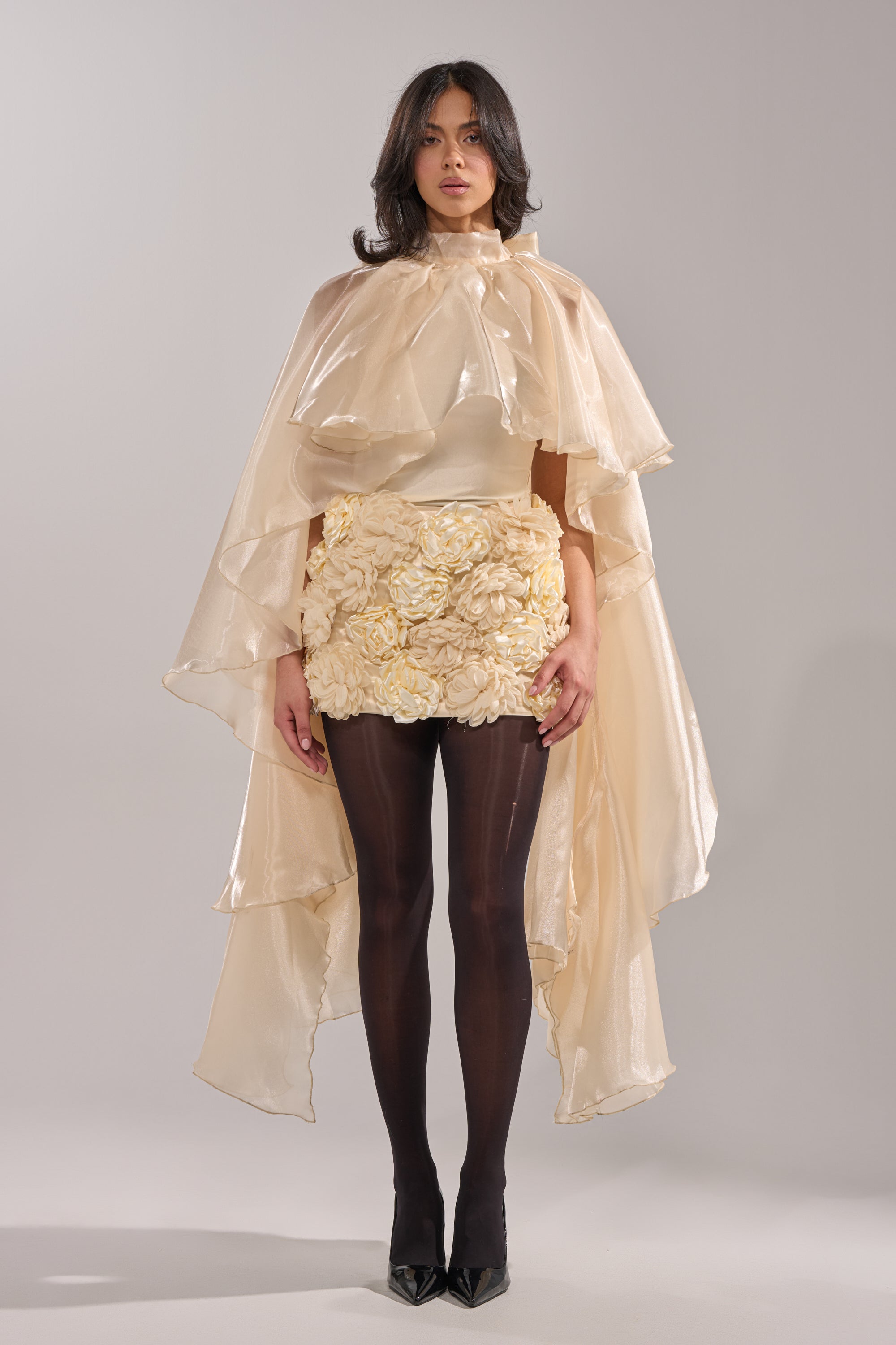 A woman wears the STAR OF THE NIGHT TOP in cream, styled with an ivory organza cape, textured floral skirt, black tights, and pointed heels against a plain light backdrop for a high-fashion look. 2