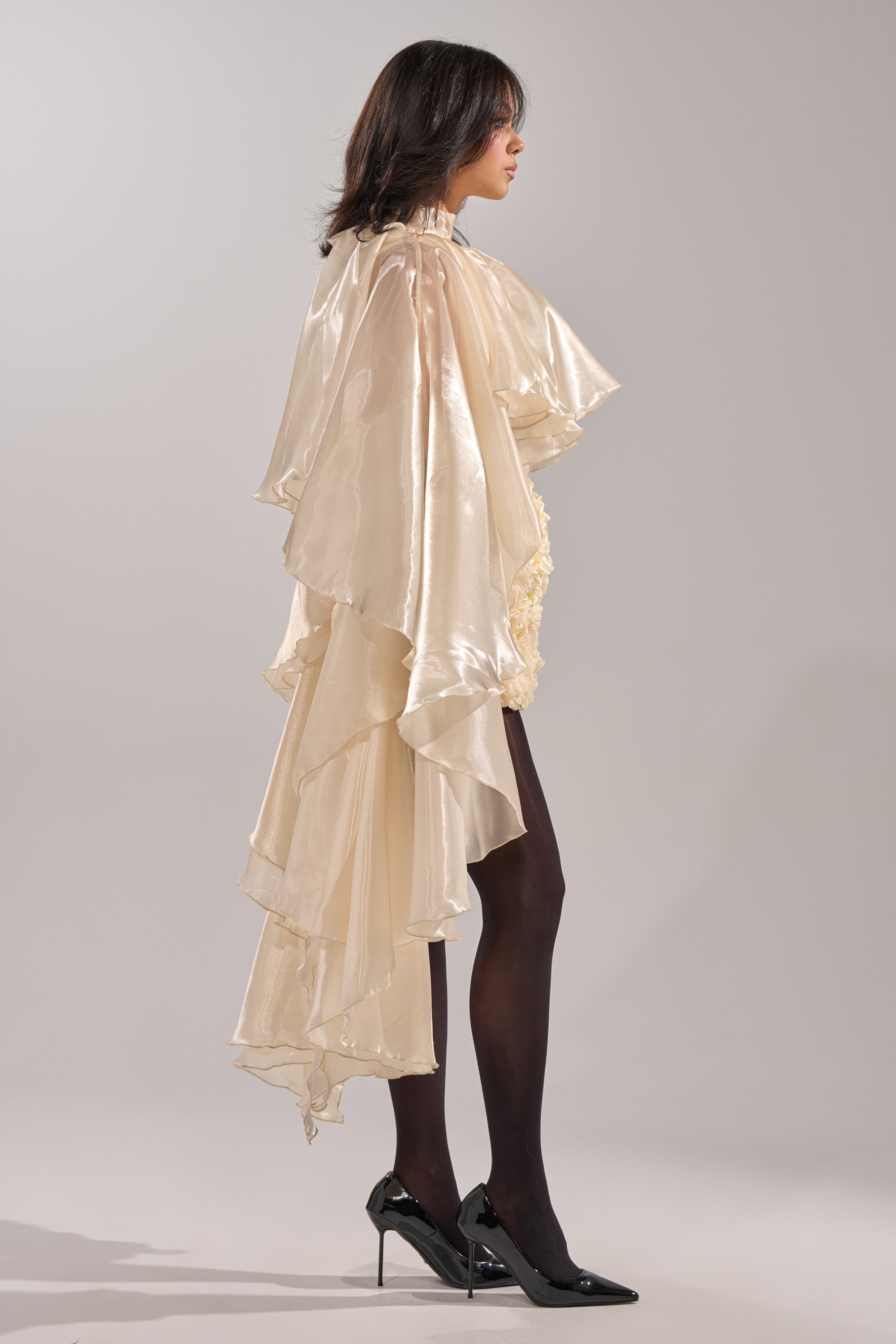 A woman in profile exudes high-fashion elegance wearing the STAR OF THE NIGHT TOP, paired with a flowing, layered cream skirt, sheer black tights, and black high heels against a plain light gray background. 3