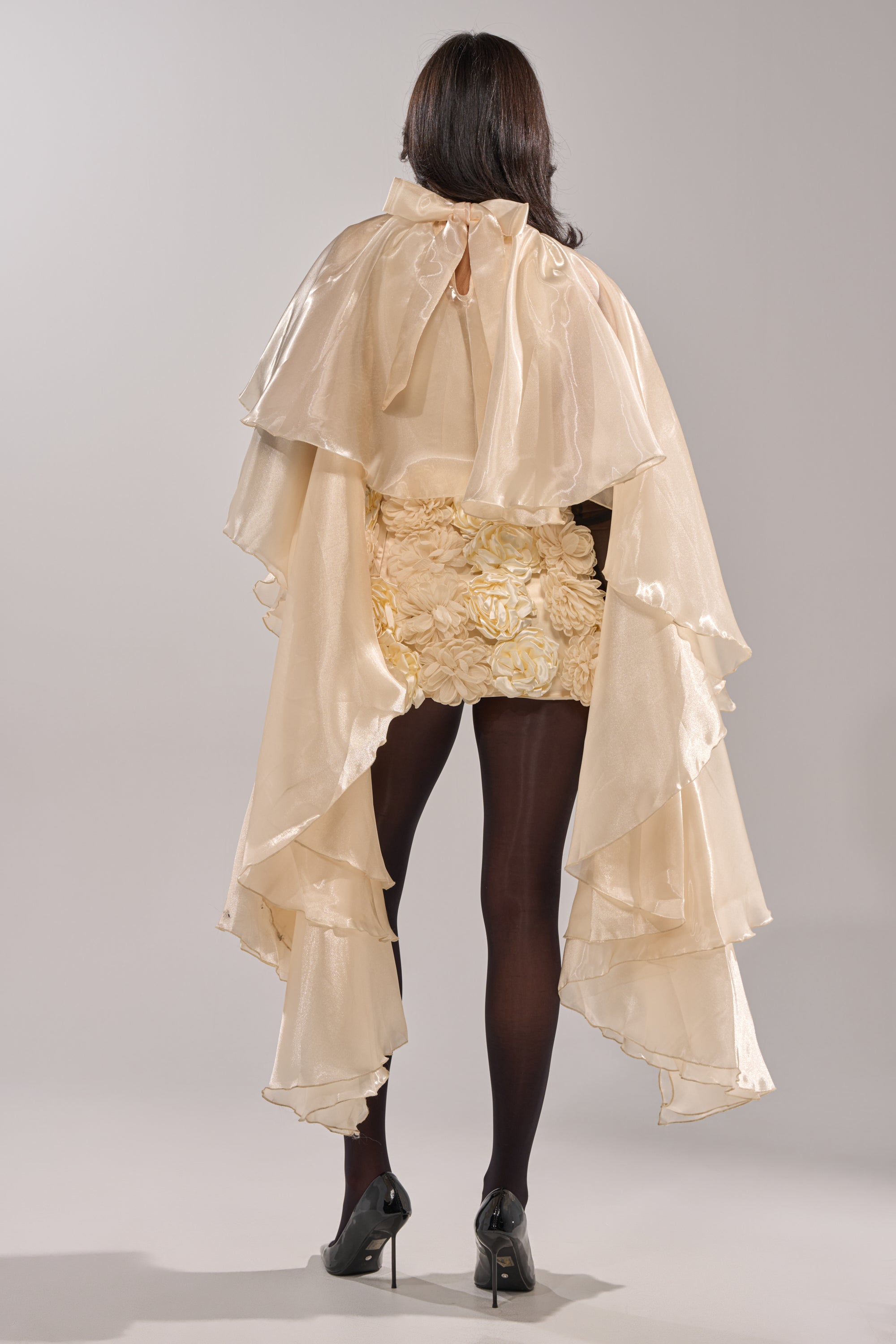 A person with long dark hair, seen from behind, models the STAR OF THE NIGHT TOP—a shiny, layered ivory organza cape and ruffled skirt—paired with black tights and heels for an elegant evening look against a plain light gray background. 4