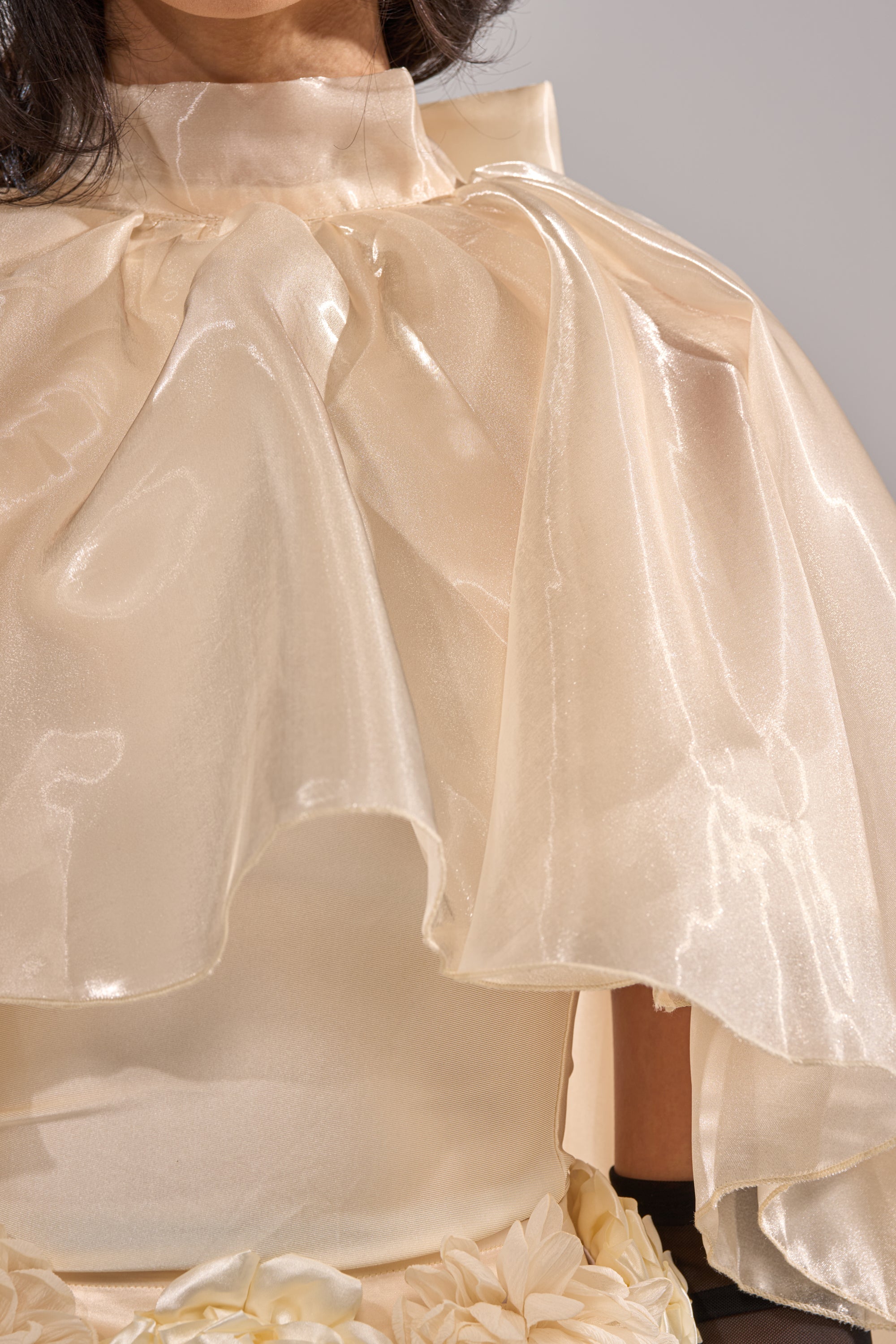 A close-up of a person wearing the STAR OF THE NIGHT TOP—a shiny, cream high-neck blouse with large flowing ruffles and floral waist details, perfect as a statement piece for elegant eveningwear. 5