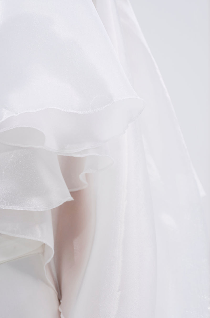 Close-up of sheer, layered white fabric with soft folds and a subtle sheen, showcasing the delicate tiered ruffles of the JUMP AROUND BLOUSE against a light background. 9