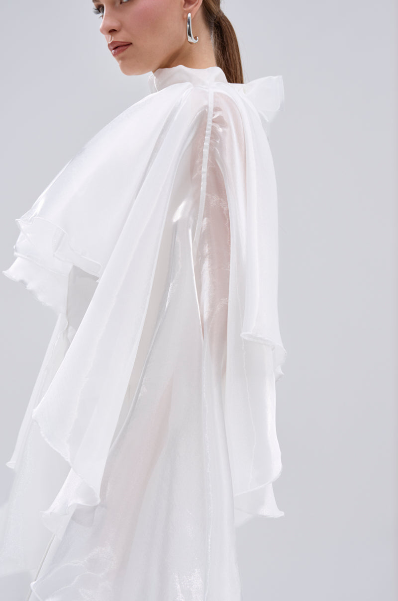 A woman with straight brown hair in a ponytail wears the JUMP AROUND BLOUSE, a sheer satin top featuring tiered ruffles and flowing sleeves, styled with modern silver earrings against a light gray background. 10