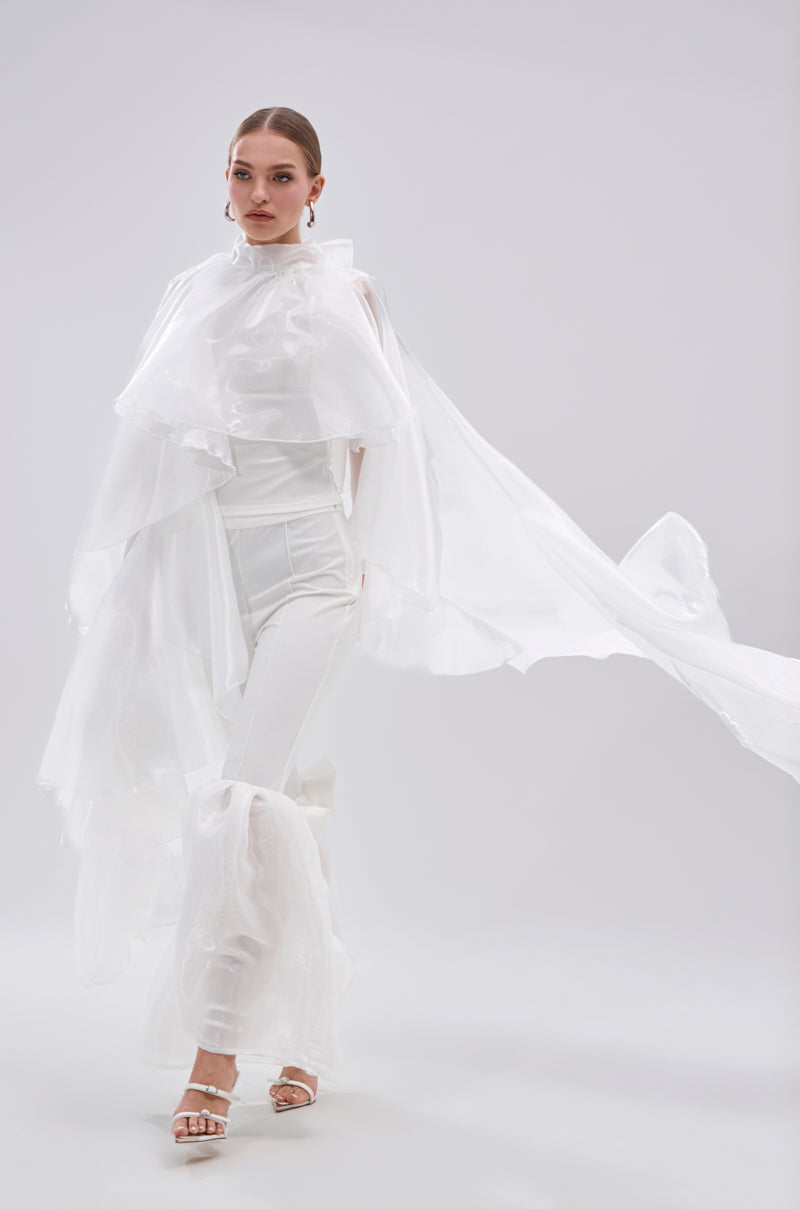 A woman stands confidently in an all-white look, featuring the JUMP AROUND BLOUSE with sheer satin and tiered ruffles, paired with white pants, heeled sandals, and statement earrings against a light backdrop. 0