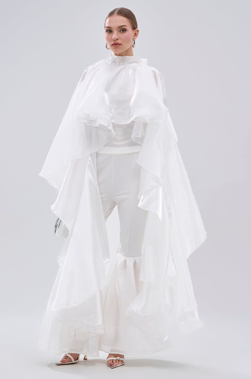A woman wears the JUMP AROUND BLOUSE, a dramatic all-white top with flowing, sheer satin fabric and wide, flared pants. Her hair is pulled back and she pairs the look with white heeled sandals against a plain light background. 1