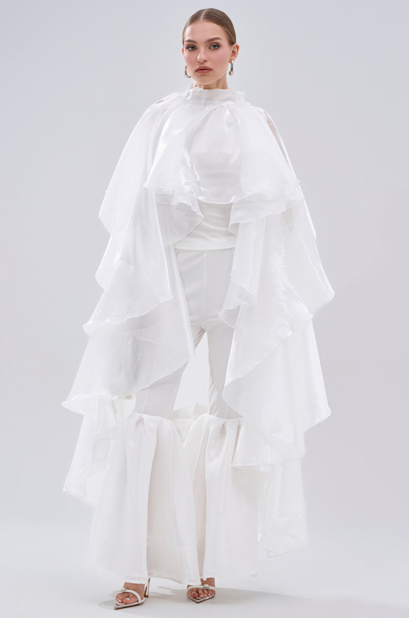 A woman models the dramatic all-white JUMP AROUND BLOUSE, with tiered ruffle satin layers cascading from shoulders to knees over flared pants. She pairs it with heels and statement earrings, set against a plain background. 2
