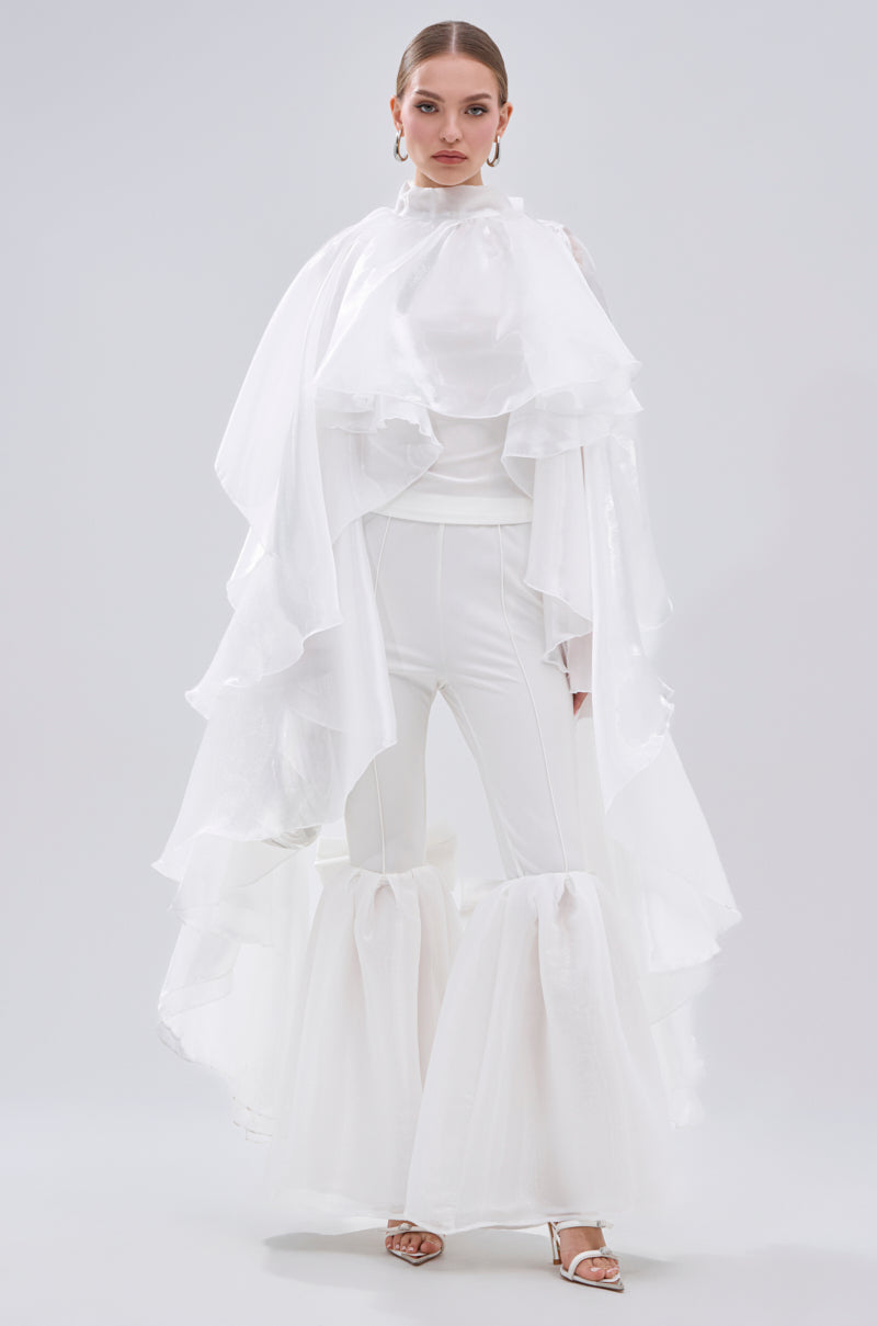 A woman wears the JUMP AROUND BLOUSE, a dramatic all-white top with tiered ruffles on the shoulders and cascading layers, styled with wide-legged pants with sheer knee ruffles and white heeled sandals. 3