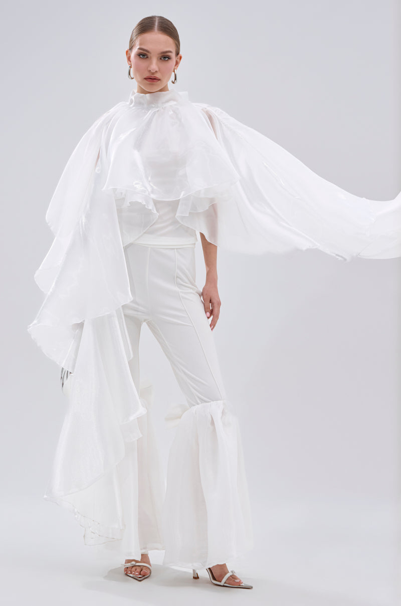 A woman confidently poses in heels and flowing pants, wearing the dramatic all-white JUMP AROUND BLOUSE featuring sheer satin and tiered ruffle sleeves, set against a plain light background. 4