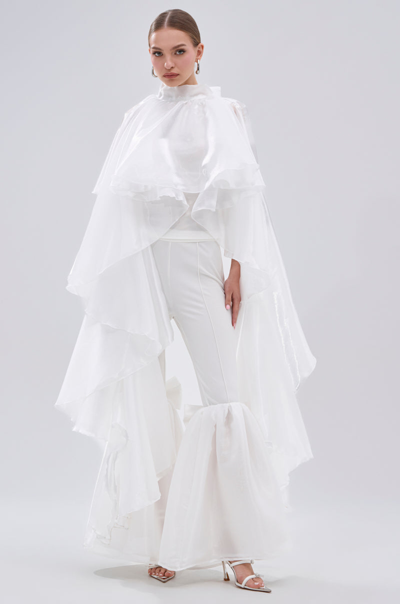 A woman models the JUMP AROUND BLOUSE, an all-white top with sheer satin tiered ruffle sleeves, paired with matching pants and white heeled sandals. She completes the look with statement earrings against a light gray background. 5
