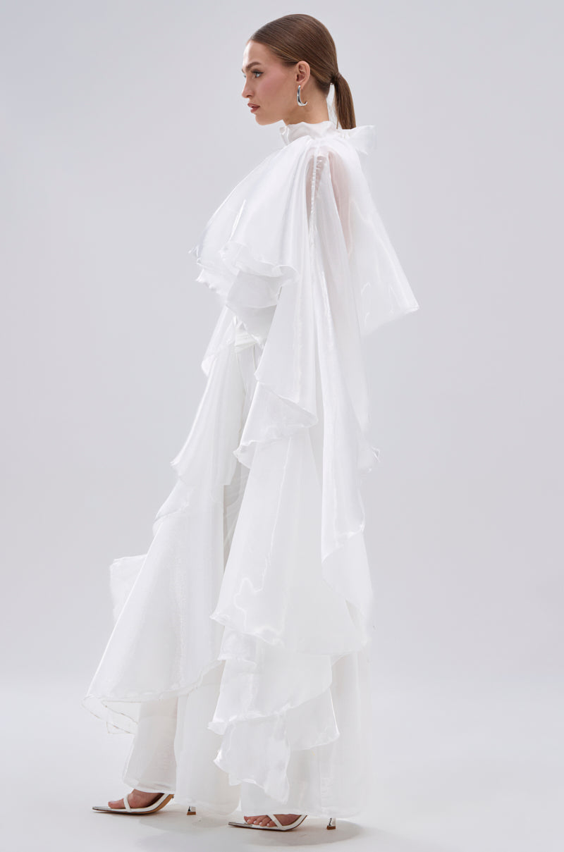 A woman in profile wears the JUMP AROUND BLOUSE—a flowing, sheer satin white top with long sleeves and tiered ruffle details—paired with white heeled sandals, against a plain light grey background. 6