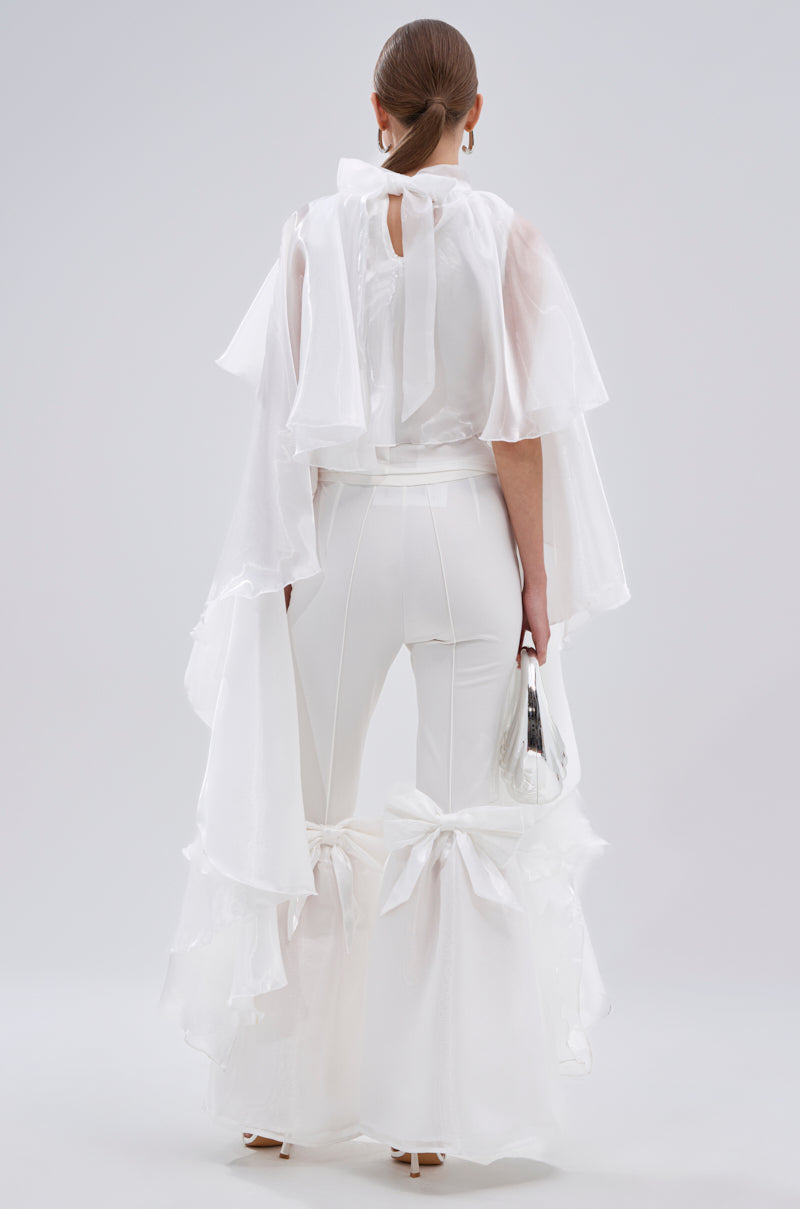A person stands facing away, wearing the JUMP AROUND BLOUSE—an all-white sheer satin top with wide sleeves and dramatic tiered ruffle details, styled with bows at the calves. They carry a small white handbag and have their hair neatly styled back. 7