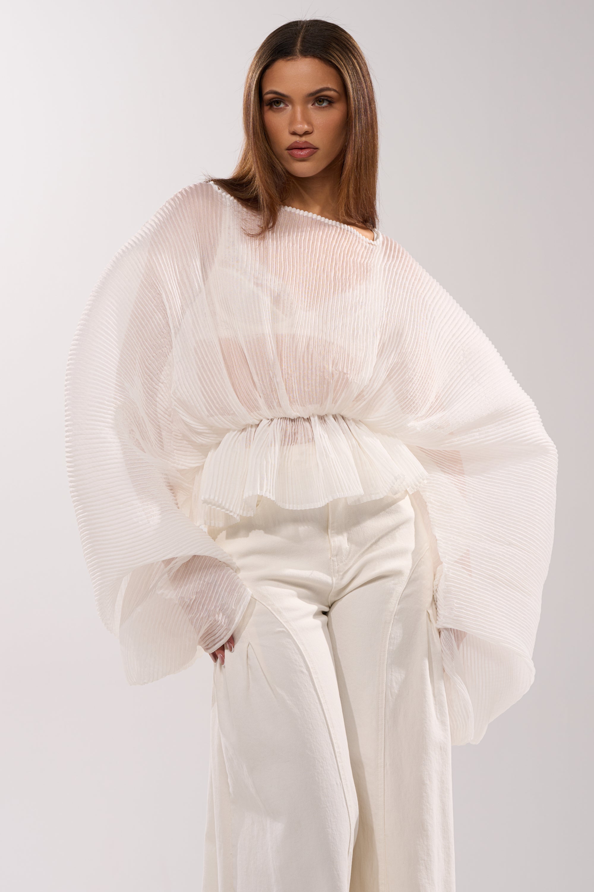 A woman in the PRETTY GIRL PROBLEMS BLOUSE IN WHITE—sheer, pleated chiffon with voluminous sleeves—pairs it with high-waisted white pants and poses confidently against a plain backdrop.