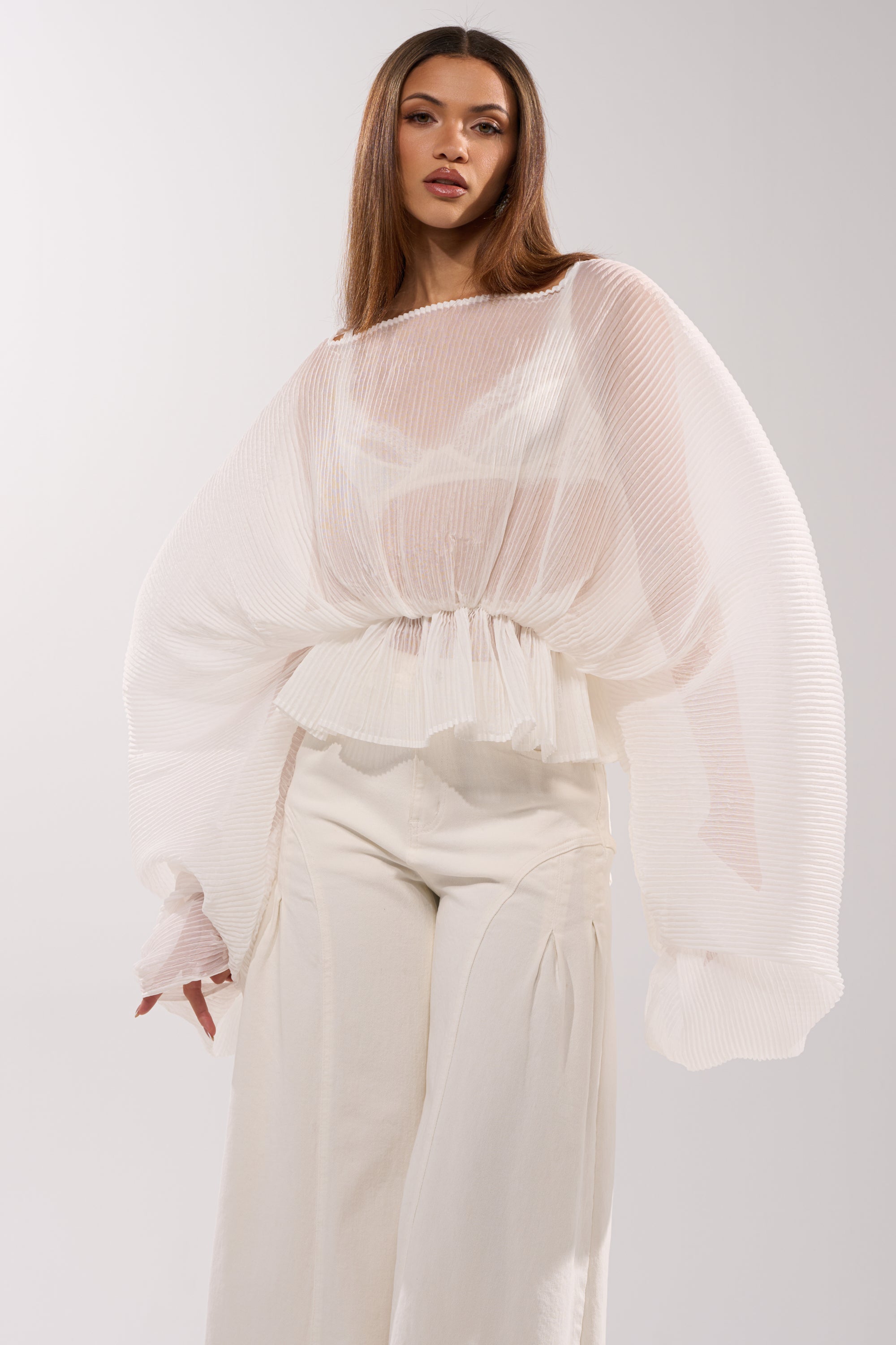 A woman wears the PRETTY GIRL PROBLEMS BLOUSE IN WHITE, featuring sheer, pleated chiffon with oversized sleeves and a peplum hem, styled with high-waisted wide-leg white pants against a plain light background.