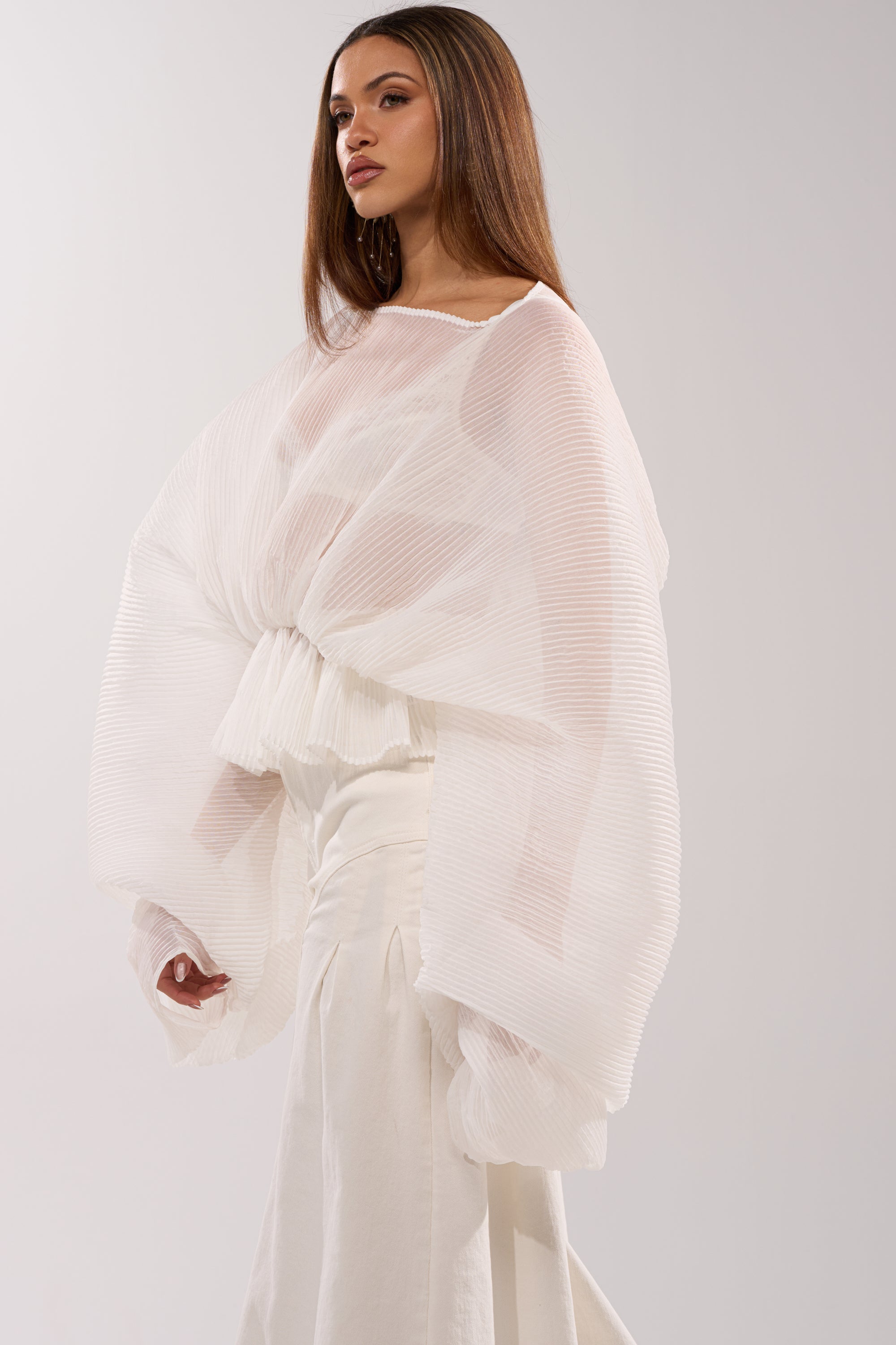 Wearing the PRETTY GIRL PROBLEMS BLOUSE IN WHITE, a woman stands against a plain, light background in a sheer, pleated chiffon blouse with voluminous sleeves, paired with a high-waisted white skirt, looking to the side.