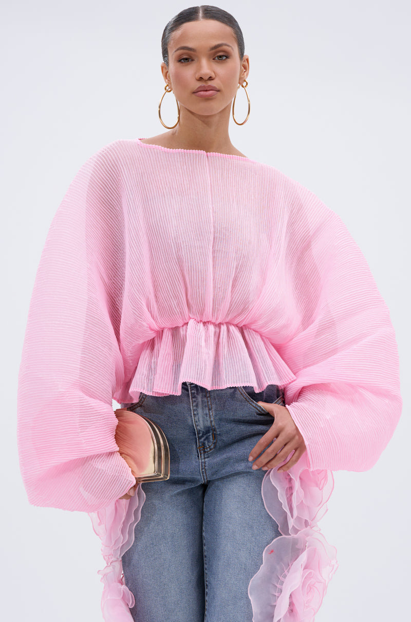 A woman poses confidently in the PRETTY GIRL PROBLEMS BLOUSE—a sheer, light pink pleated top with voluminous batwing sleeves and ruffled details—paired with blue jeans, large hoop earrings, and a blush clutch.