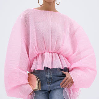A woman poses confidently in the PRETTY GIRL PROBLEMS BLOUSE—a sheer, light pink pleated top with voluminous batwing sleeves and ruffled details—paired with blue jeans, large hoop earrings, and a blush clutch.