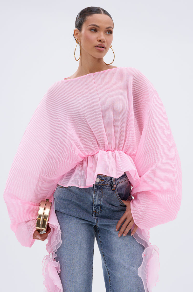 A woman wears the PRETTY GIRL PROBLEMS BLOUSE—a sheer, pleated pink top with batwing sleeves—styled with blue jeans, a gold clutch, and large hoop earrings. Her hair is tied back for a chic look.