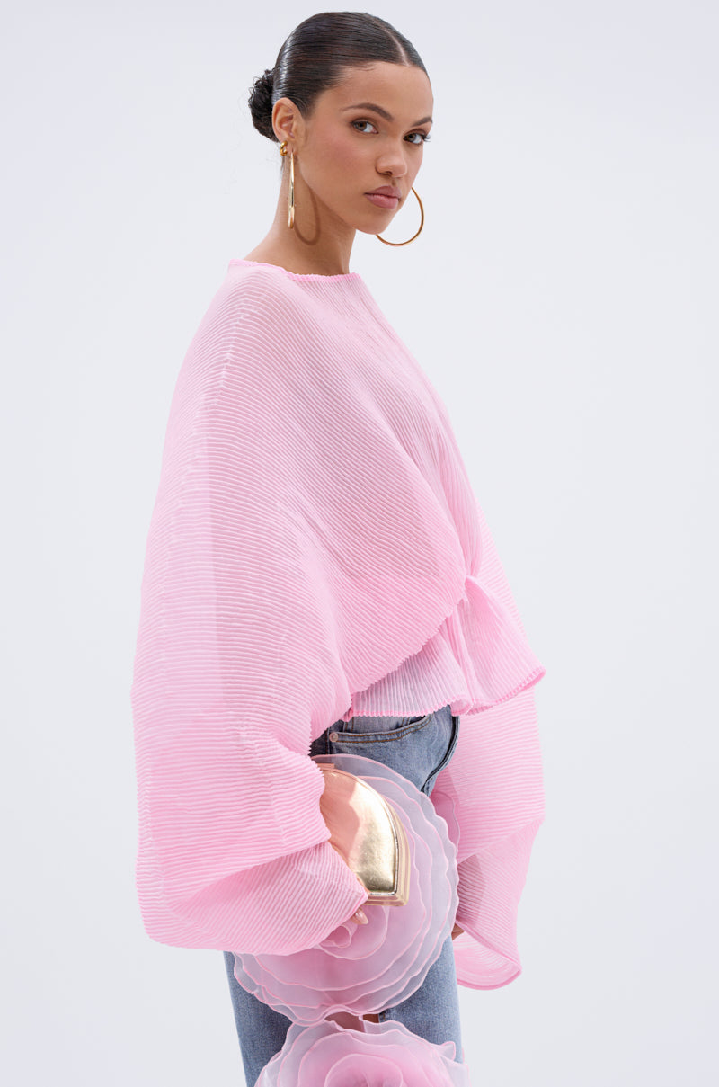 A woman with slicked-back hair wears the PRETTY GIRL PROBLEMS BLOUSE—a loose, textured pink top with batwing sleeves—styled with blue jeans, hoop earrings, and a metallic floral clutch against a plain light background.