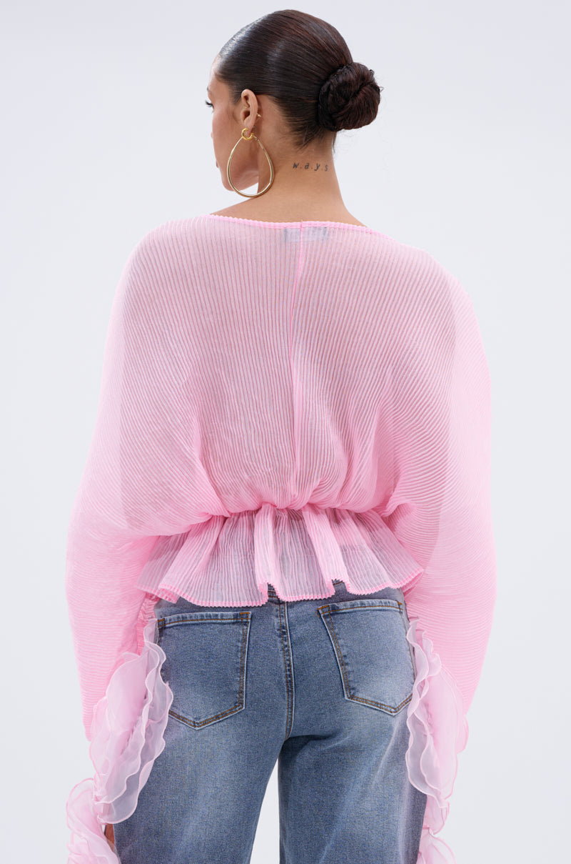 A woman with a neat bun and large hoops wears the PRETTY GIRL PROBLEMS BLOUSE—a sheer pink, pleated top with ruffled sleeves—paired with blue jeans, seen from behind against a plain light background.