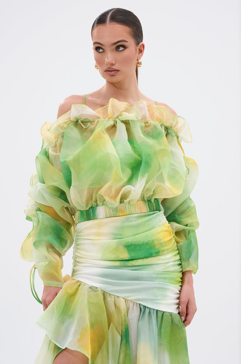Wearing the LIMONE CHIFFON BLOUSE with an off-the-shoulder neckline and sheer watercolor patterns, paired with a matching ruched skirt, she styles slicked-back hair, gold hoops, and looks to the side. 0